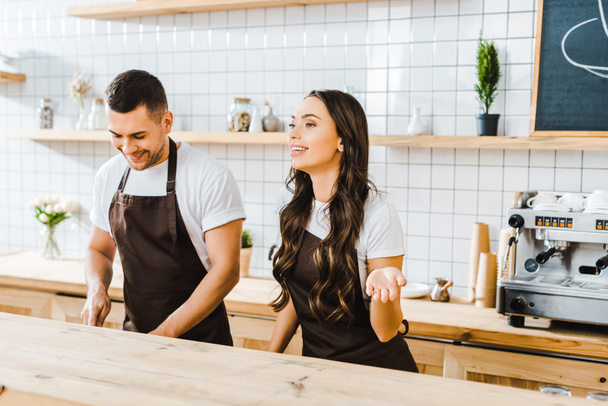 attractive cashier standing behind wooden bar counter and smiling wile barista working in coffee house - Photo, Image