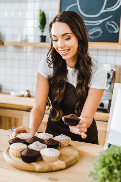 selective focus of attractive waitress standing behind bar counter and putting cupcakes on wooden tray in coffee house - Zdjęcie, obraz