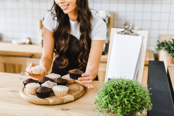 vista cortada de garçonete atraente colocando cupcakes em bandeja de madeira na casa de café
 - Foto, Imagem