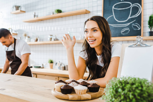 selective focus of attractive brunette waitress standing behind bar counter with cupcakes and waving wile cashier working in coffee house - Photo, Image