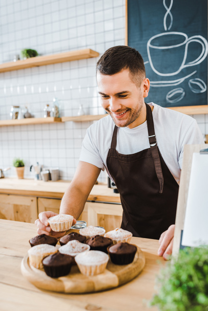 handsome waiter standing behind bar counter and putting cupcakes on wooden tray in coffee house - Photo, Image