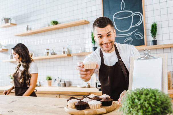 selectieve aandacht van knappe barista geven cupcake wile aantrekkelijke brunette kassier werken in koffiehuis - Foto, afbeelding