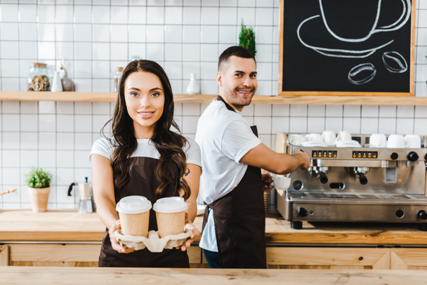 brünette Kassiererin steht und hält Pappbecher mit einem Mann, der Kaffee im Kaffeehaus kocht - Foto, Bild