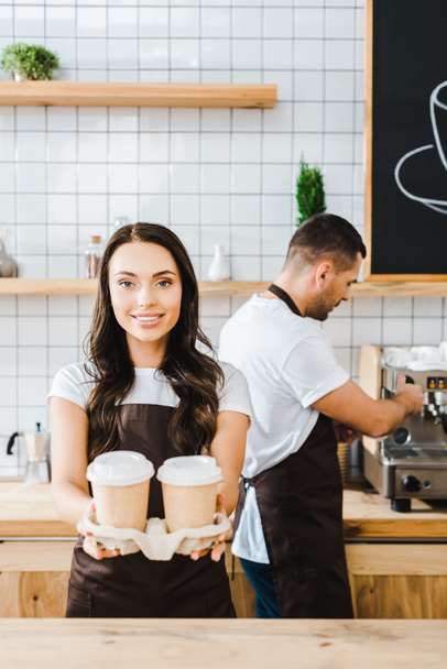 brunette attractive cashier standing and holding paper cups wile barista working in coffee house - Photo, Image