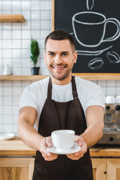 selective focus of handsome barista in brown apron holding cup and saucer in coffee house - Photo, Image