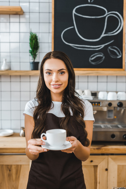 attractive barista in brown apron holding cup and saucer in coffee house - Фото, изображение