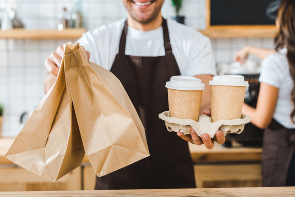 vista recortada de cajero sosteniendo vasos de papel y bolsas barista morena astuta haciendo café en la cafetería
 - Foto, Imagen