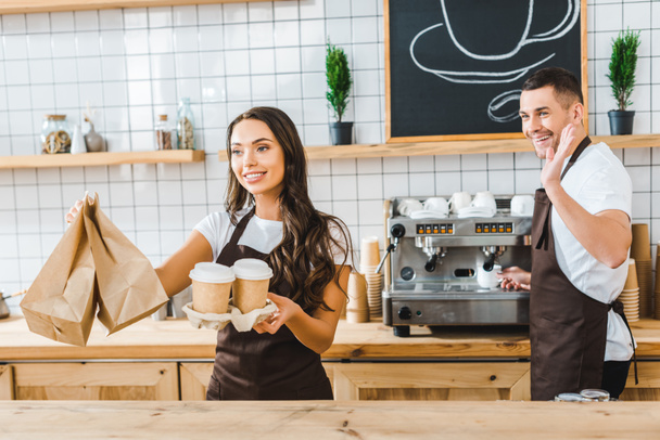 attractive brunette cashier holding paper cups and bags wile barista waving in coffee house - Photo, Image
