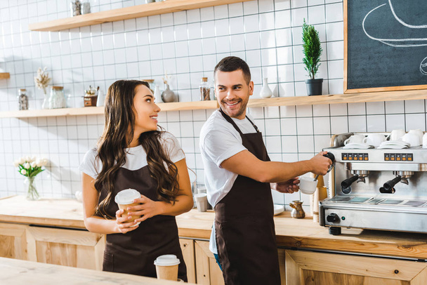 handsome barista making coffee and attractive brunette cashier holding paper cup in coffee house - Photo, Image