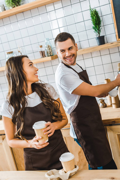 barista in brown apron making coffee and attractive brunette cashier holding paper cup in coffee house - Photo, Image
