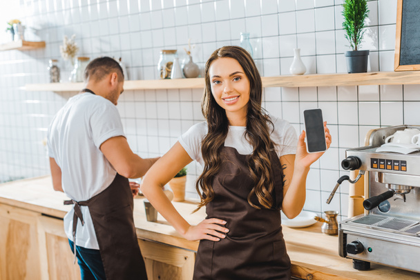 attraente bruna cassiere mostrando smartphone con schermo bianco wile barista che lavora in caffetteria
 - Foto, immagini