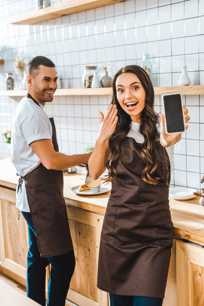 brunette cashier showing smartphone with blank screen wile barista smiling in coffee house - Photo, Image