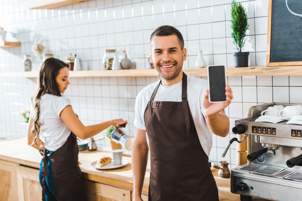 hübsche Kassiererin zeigt Smartphone mit leerem Bildschirm wile brünette Barista arbeitet im Kaffeehaus - Foto, Bild