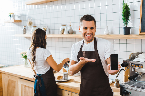 handsome cashier pointing with hand to smartphone with blank screen wile barista working in coffee house - Photo, Image