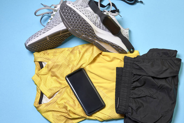 sapatos de corrida com um par de shorts, uma camisa amarela e acessórios tecnológicos em um fundo azul
 - Foto, Imagem