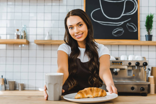 attractive brunette cashier standing behind bar counter with cup, plate and croissant in coffee house - Photo, Image