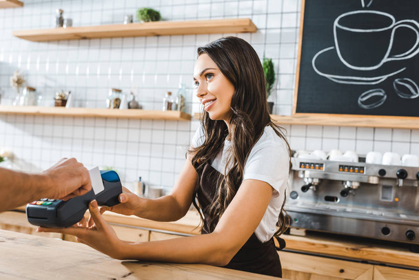attraktive Kassiererin steht in brauner Schürze neben Theke und hält Terminal wile Mann zahlt mit Kreditkarte im Kaffeehaus - Foto, Bild