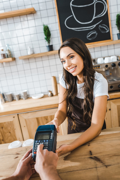 attractive cashier standing near bar counter in brown apron and holding terminal wile man making transaction in coffee house - Photo, Image
