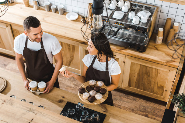 attractive cashier with cupcakes and handsome barista with paper cups standing behind bar counter in coffee house - Photo, Image