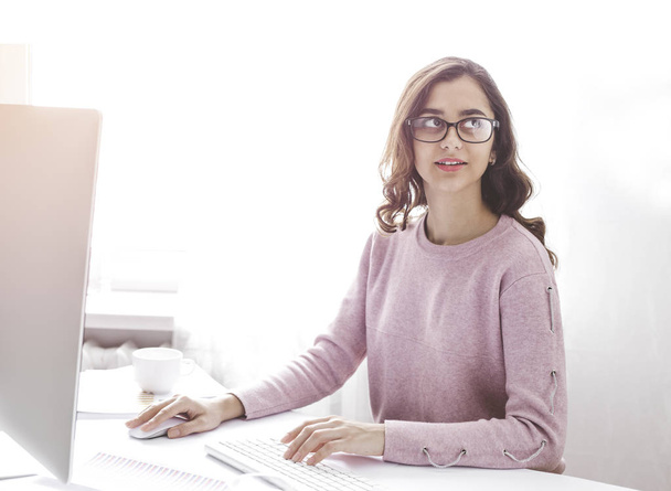 Bilgisayarda çalışan ve müşterisiyle telefonda konuşan gözlüklü mutlu Hintli kız. Genç, gülümseyen esmer kadın işyerindeki parlak, modern bir ofiste oturuyor.. - Fotoğraf, Görsel