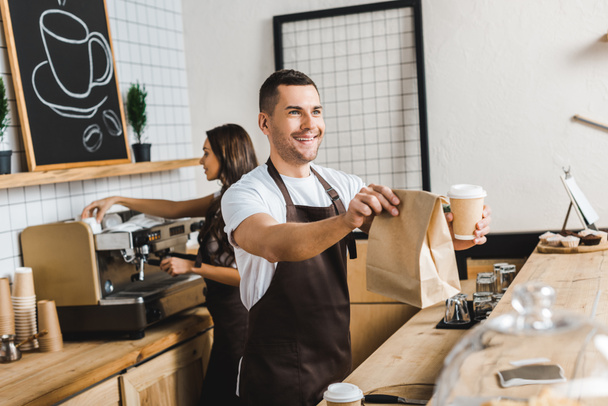 apuesto cajero dando taza de papel y bolsa barista astuto haciendo café detrás del mostrador de la barra en la cafetería
 - Foto, imagen