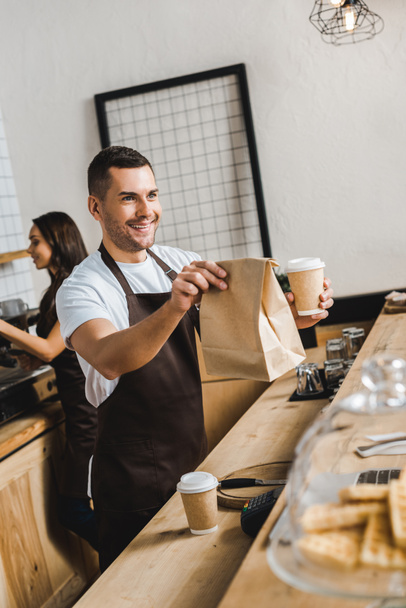 bonito caixa em avental marrom dando copo de papel e saco wile barista trabalhando atrás bar contador na casa de café
 - Foto, Imagem