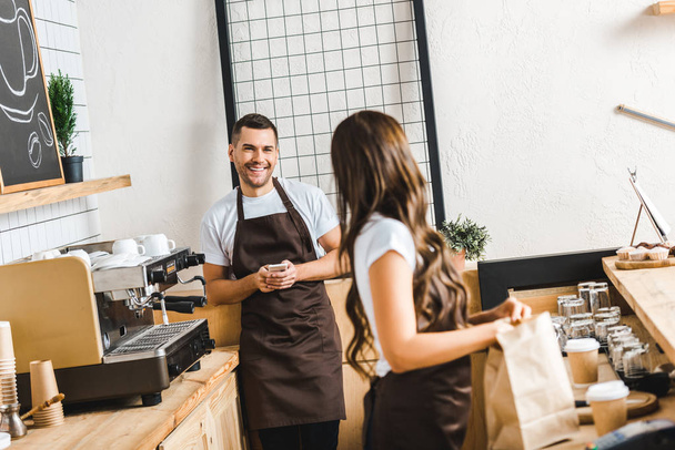 селективное внимание привлекательного кассира со смартфоном и баристой, стоящей за барной стойкой в кофейне
 - Фото, изображение