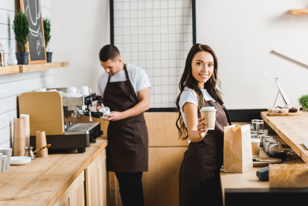 atractiva cajera sonriente con taza de papel con barista en delantal marrón haciendo café detrás del mostrador de la barra en la cafetería
 - Foto, Imagen