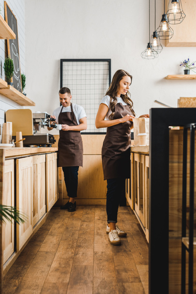 attractive cashier with paper bag and barista in brown apron making coffee behind bar counter in coffee house - Photo, Image