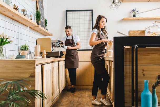 attractive cashier with paper bag and barista making coffee behind bar counter in coffee house - Photo, Image