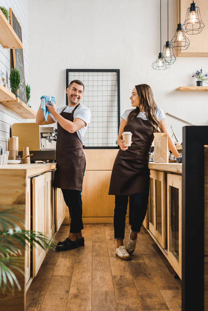 barista and cashier in aprons standing and talking behind bar counter in coffee house - Photo, Image