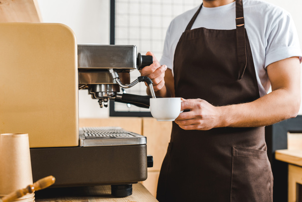 vista recortada de barista guapo sosteniendo la taza y haciendo café con la máquina en la cafetería
 - Foto, Imagen