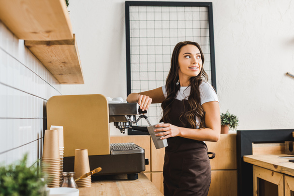 attractive brunette barista holding cup and making coffee with machine in coffee house - Photo, Image