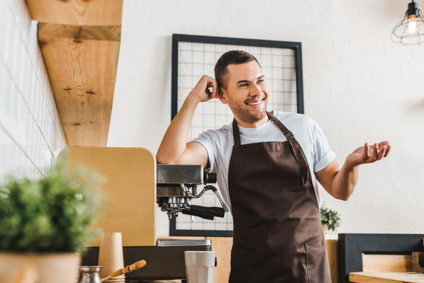 smiling handsome barista in brown apron standing near coffee machine in coffee house - Фото, зображення