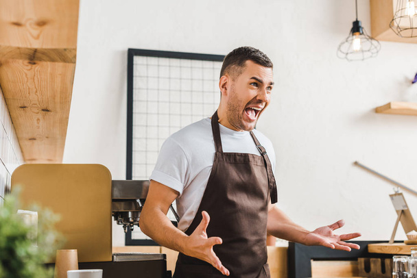 agressivo bonito barista no avental marrom em pé perto da máquina de café na casa de café
 - Foto, Imagem