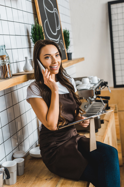 cashier in brown apron talking on smartphone, holding clipboard and sitting on bar counter in coffee house - Photo, Image