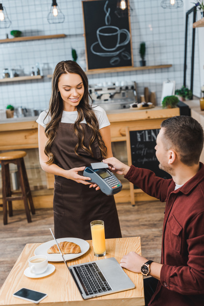 attractive waitress holding terminal wile handsome man sitting at table with laptop and paying with credit card in coffee house - Photo, Image