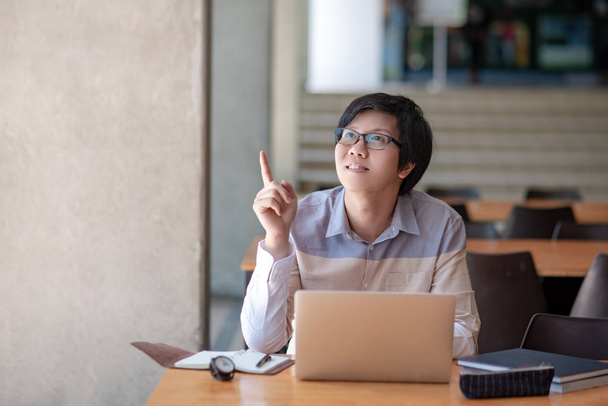 Joven estudiante universitario asiático con gafas apuntando con el dedo hacia arriba mientras usa la computadora portátil en la biblioteca de la universidad. Oportunidad de educación o beca. Conceptos de ideas e inspiración
 - Foto, Imagen