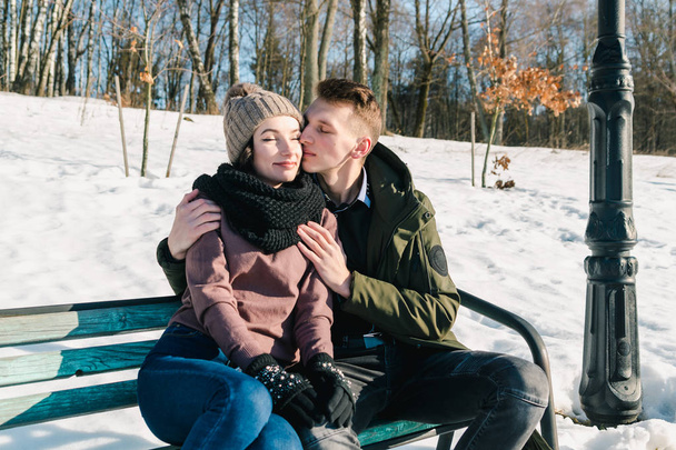 Bella giovane coppia innamorata seduta su una panchina del parco in una chiara giornata invernale soleggiata. Ragazzo abbraccia e bacia la sua ragazza
 - Foto, immagini