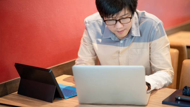 Αρσενικό Ασίας επιχειρηματίας φορώντας γυαλιά εργασία με σύγχρονο φορητό υπολογιστή και ψηφιακό tablet σε ξύλινο τραπέζι στο καφενείο. Εργαστείτε έξυπνα στην ψηφιακή πλατφόρμα. Διαδίκτυο των πραγμάτων (Iot) έννοια - Φωτογραφία, εικόνα