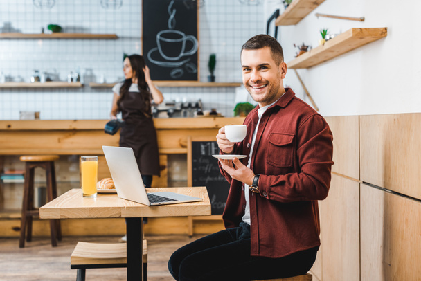 handsome freelancer sitting at table with laptop, drinking coffee and smiling wile waitress standing near bar in coffee house - Photo, Image