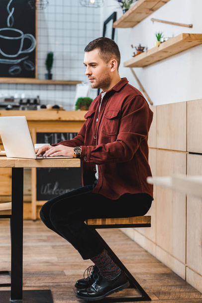 handsome freelancer in burgundy shirt sitting at table with laptop and typing in coffee house - Photo, Image