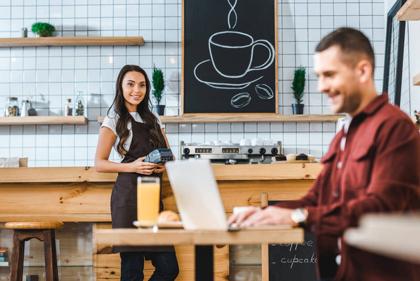 selective focus of waitress standing near bar counter with terminal wile freelancer in burgundy shirt sitting at table in coffee house - Photo, Image