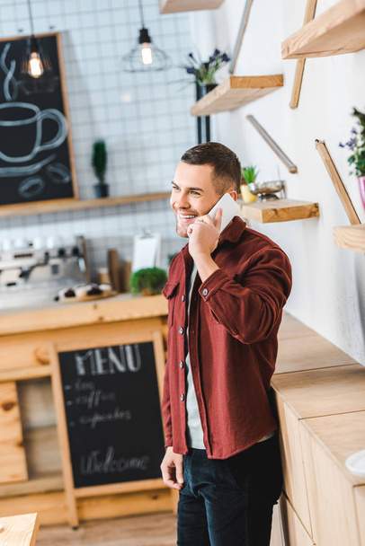 hombre guapo en camisa de color burdeos y jeans de pie y hablando en el teléfono inteligente en la cafetería
 - Foto, imagen
