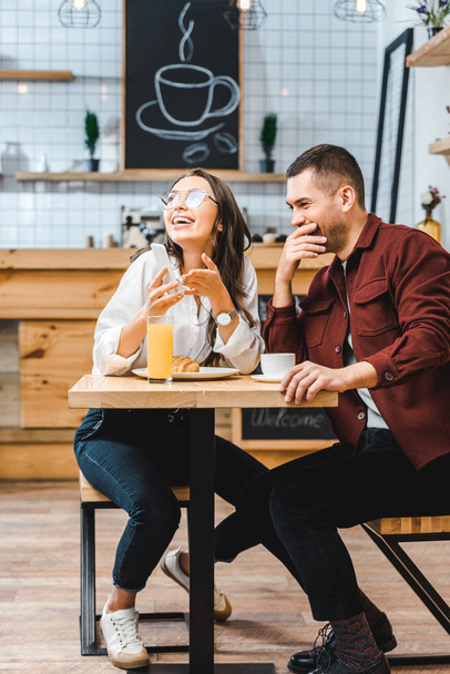 attractive brunette woman and handsome man in burgundy shirt sitting at table, laughing and looking to smartphone in coffee house - Photo, Image