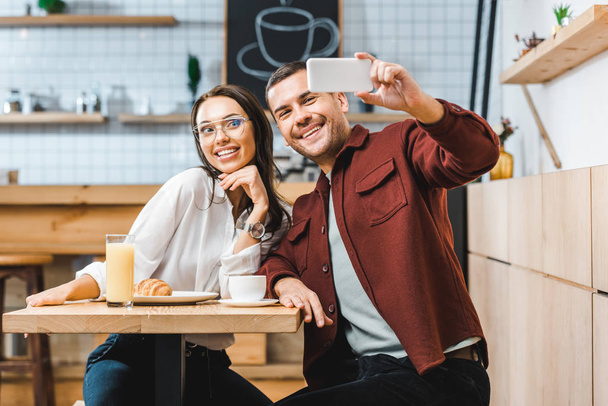 attractive brunette woman and handsome man in burgundy shirt sitting at table and taking selfie to smartphone in coffee house - Photo, Image