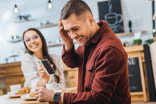 Selektivní fokus pohledný muž v vínové tričko sedí u stolu a se smíchem wile atraktivní bruneta žena zobrazeno smartphone s prázdnou obrazovkou v kavárně - Fotografie, Obrázek