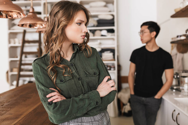 Upset woman with wavy hair holding hands together sadly looking aside with asian man on background. Young international couple quarreling spending time on kitchen at home - Photo, Image