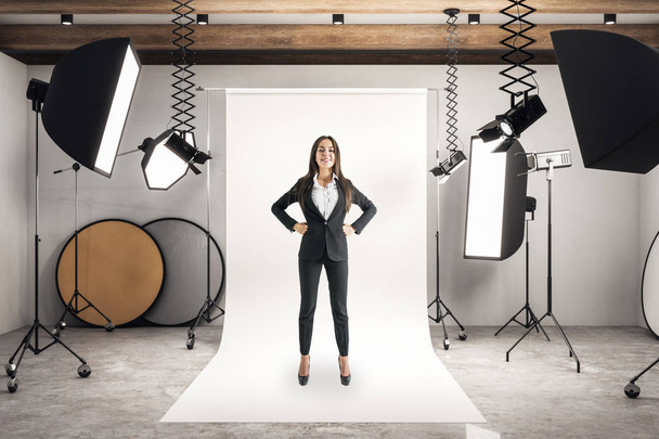 Happy young businesswoman standing in photo studio with daylight and professional equipment. - Фото, изображение