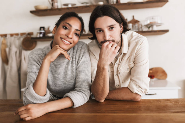 Attractive man and pretty smiling african american woman leaning on table happily looking in camera together. Young international couple dreamily spending time together in cozy kitchen at home - Photo, Image